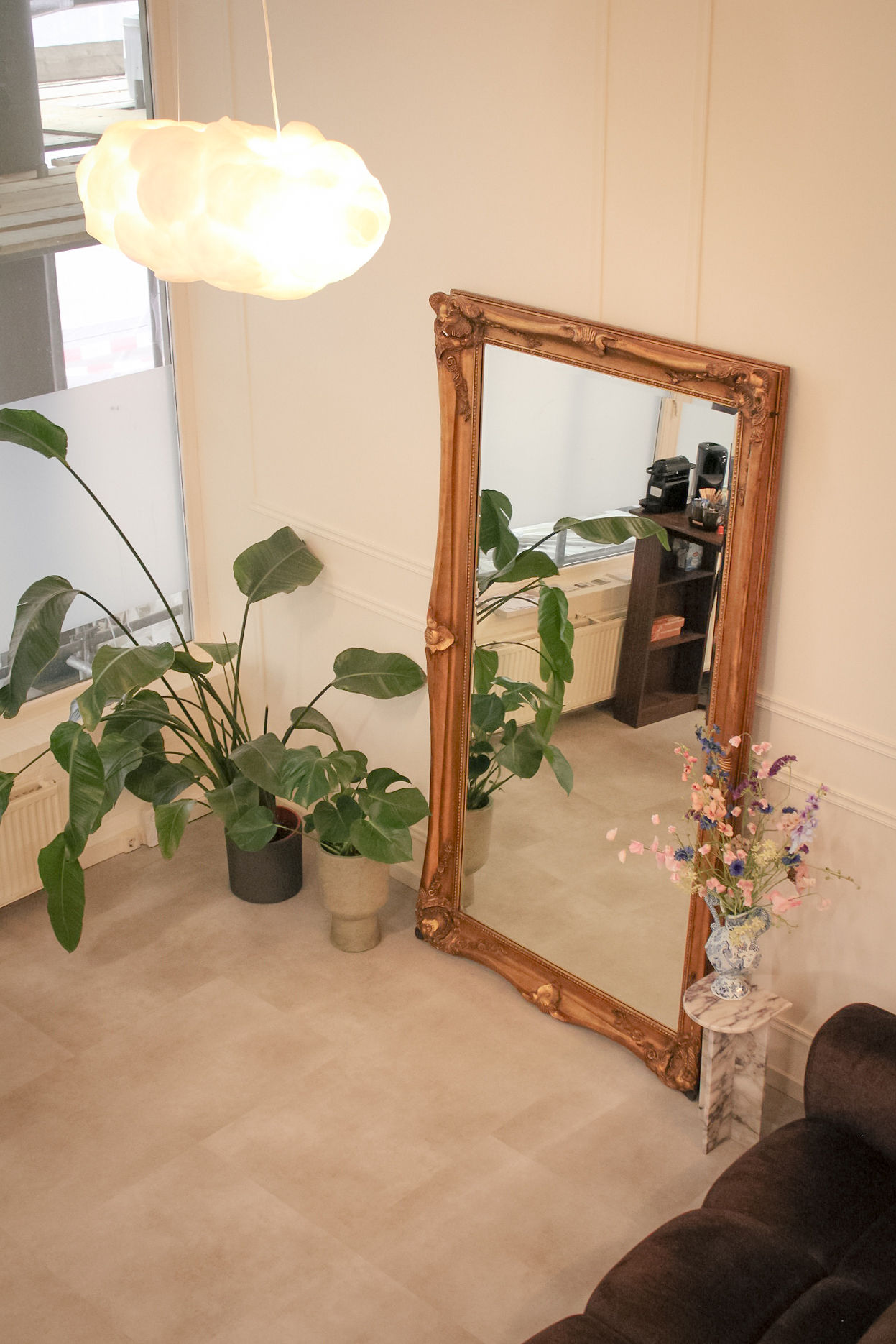De studio met een wolkenlamp aan het plafond, een spiegel in een gouden lijst, planten en een bank